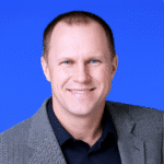 Smiling man in a gray patterned blazer and dark shirt poses in front of a solid blue background.